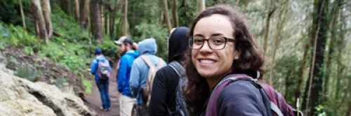 A group of people on a wooded trail, the last person on the trail is looking back and smiling