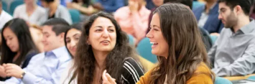 Two young women smiling and chatting in a large lecture hall